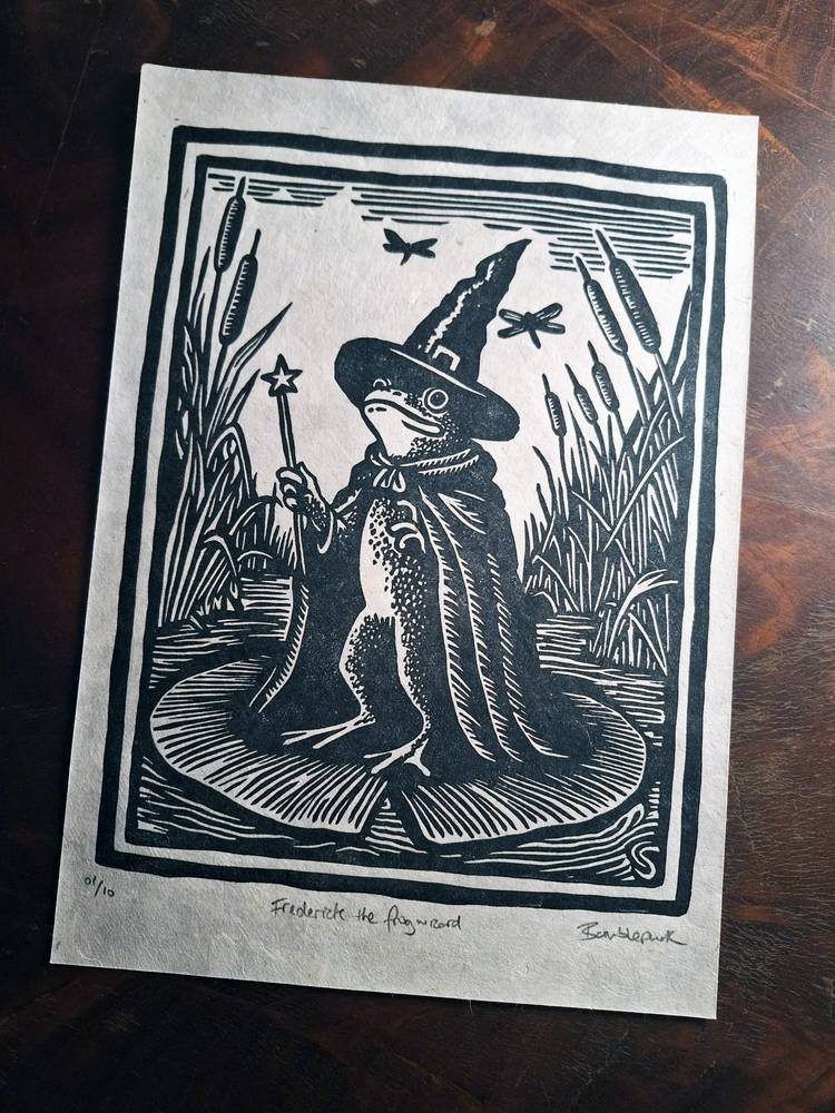Bumblepunk’s black and white linocut of Frederick the frog wizard, wearing a pointed hat and cape and holding a star-tipped wand beside reeds.
