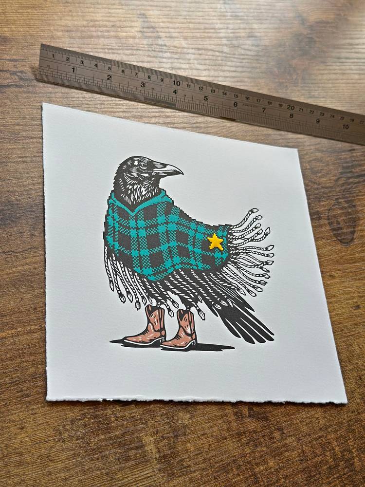 A linocut crow wears a turquoise checked poncho, yellow sheriff’s badge and brown cowboy boots on white paper.