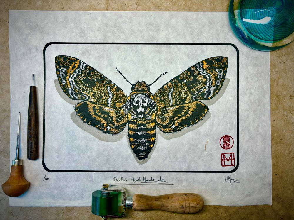 A detailed death’s-head hawk moth linocut in yellow, black and grey on textured paper, photographed with printmaking tools.