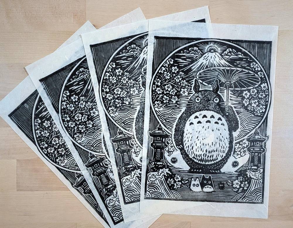 A stack of black-and-white Totoro in Bloom linocut prints by Phew What A Relief, showing Totoro with an umbrella, Mount Fuji and blossom.
